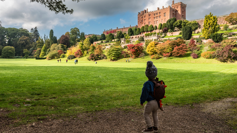 View across the sweeping lawn at Powis Castle and Garden in autumn. A young child wearing a bobble hat and backpack stands in the foreground looking towards the castle, which rises above terraced gardens and distinctive clipped yew topiary. Vibrant autumn foliage in shades of red, orange and gold brightens the hillside beneath dramatic clouds and patches of blue sky.